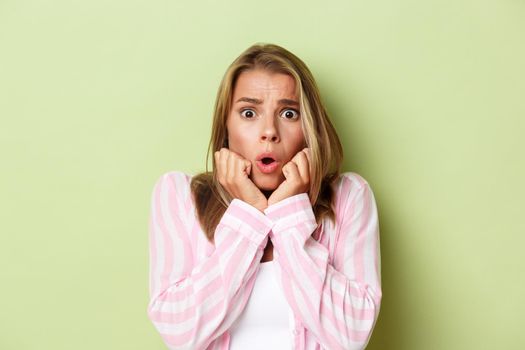 Close-up of shocked blond woman looking scared, standing ambushed with something scary over green background.