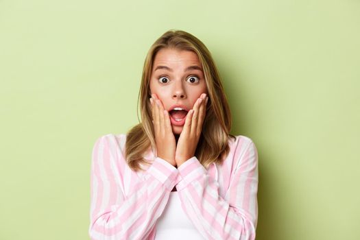 Close-up of attractive blond girl gasping, looking surprised with advertisement, standing over green background.