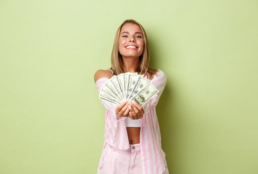 Portrait of blond attractive woman in pink outfit, giving you money and smiling, standing over green background.