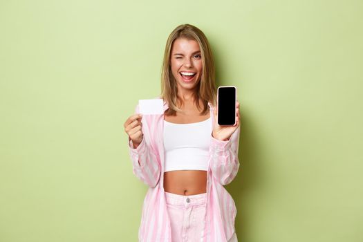 Image of sassy modern girl with blond short hair, showing credit card and mobile phone screen, winking and smiling while advertising application, standing over green background.