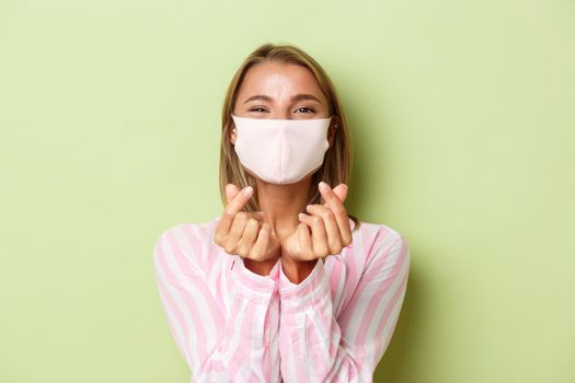 Concept of quarantine, coronavirus and lifestyle. Close-up of beautiful blond female model in face mask and pink shirt, showing heart sign, standing over green background.