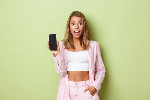 Image of blond attractive female model in pink shirt, showing smartphone screen and looking excited, standing over green background.