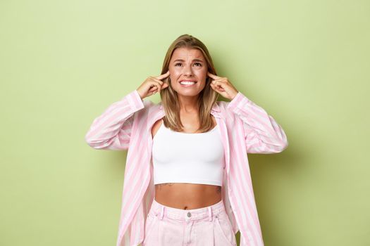 Portrait of annoyed woman complaining on loud neighbours, looking up and frowning, shut ears with fingers, standing over green background.