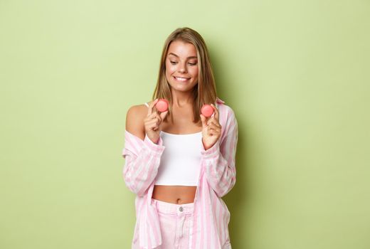 Portrait of cute glamour girl with blond hair, looking at tasty macaroons and smiling with temptation, standing over green background.