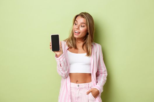 Image of blond attractive female model in pink shirt, showing smartphone screen and looking amazed, standing over green background.