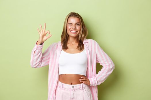 Portrait of attractive stylish woman with blond short hairstyle, wearing pink shirt with jeans, showing okay sign in approval, recommend something good, standing over green background.