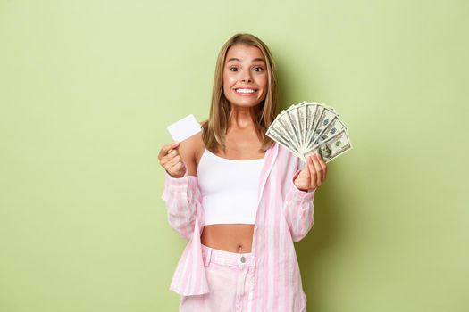 Image of successful blond woman in pink shirt, showing credit card and money, smiling happy, standing over green background.