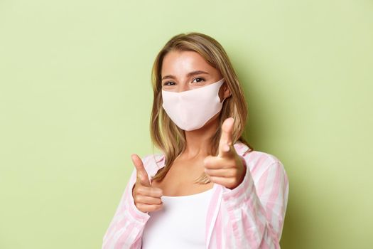 Concept of covid-19, social-distancing and lifestyle. Close-up of pretty blond girl in face mask, pointing fingers at camera, congratulating you, standing over green background.