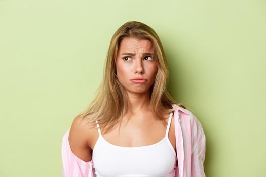 Close-up of worried blond woman in pink shirt, looking at upper left corner and sulking sad, standing over green background.