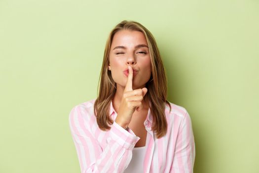 Close-up of blond flirty woman in pink shirt, hushing at camera, share a secret, standing against green background.