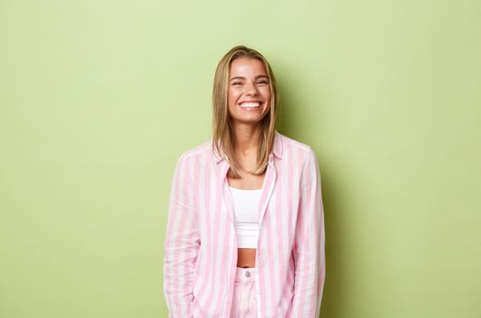 Image of cheerful blond woman with white perfect smile, standing over green background happy, wearing pink outfit.