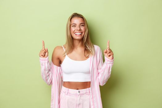 Happy beautiful blond woman in pink outfit , laughing and smiling with white teeth, pointing fingers up, showing logo, standing against green background.