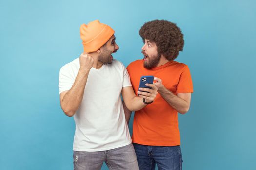 Portrait of two excited amazed young adult hipster men standing with cell phone and celebrating success, clenched fists, looking at each other. Indoor studio shot isolated on blue background.
