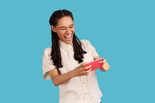 Emotional happy woman with black dreadlocks stares at smartphone camera, plays games, yelling happily while gaming smiles excited, wearing white shirt. Indoor studio shot isolated on blue background.