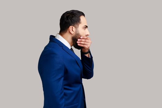 Side view of attractive bearded businessman closing mouth with hand, afraid to say too much, embarrassment, wearing official style suit. Indoor studio shot isolated on gray background.