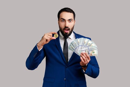 Shocked man showing dollar bills and golden coin of crypto currency, transferring money into digital bitcoins, wearing official style suit. Indoor studio shot isolated on gray background.