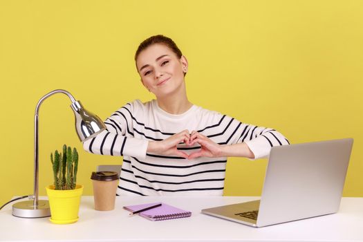 Portrait of satisfied pretty woman with kind smile sitting at workplace, showing romantic heart shape with hand, expressing love. Indoor studio studio shot isolated on yellow background.