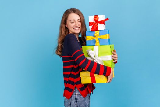 Attractive woman wearing striped casual style sweater, keeping eyes closed while embracing many present boxes, extremely happy to celebrate birthday. Indoor studio shot isolated on blue background.