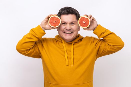 Portrait of smiling middle aged man having fun, holding two halves of grapefruit, expressing positive emotions, wearing urban style hoodie. Indoor studio shot isolated on white background.