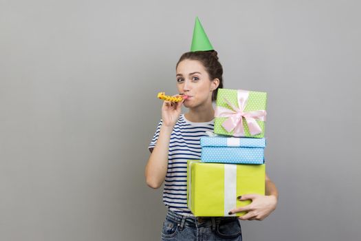 Portrait of happy delighted woman wearing striped T-shirt and party cone, holding stack of present boxes and blowing party horn. Indoor studio shot isolated on gray background.