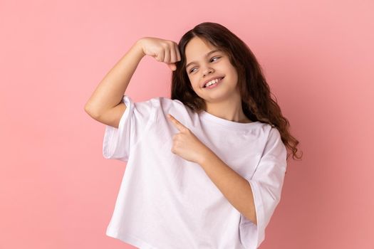 Portrait of smiling little girl wearing white T-shirt showing her arm muscle and point it with finger, proud of her strength and leadership skills. Indoor studio shot isolated on pink background.