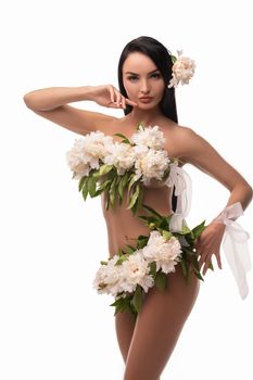 Self assured young naked female model with long dark hair covering body with bunch of fresh delicate flowers and looking at camera against white background