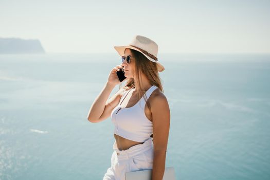 Successful business woman in yellow hat working on laptop by the sea. Pretty lady typing on computer at summer day outdoors. Freelance, travel and holidays concept.