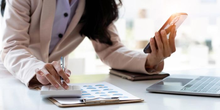 Business woman or accountant hand using calculator, working on mobile smart phone and laptop computer with documentary data graph paper on the table at modern home office. Online marketing concept..