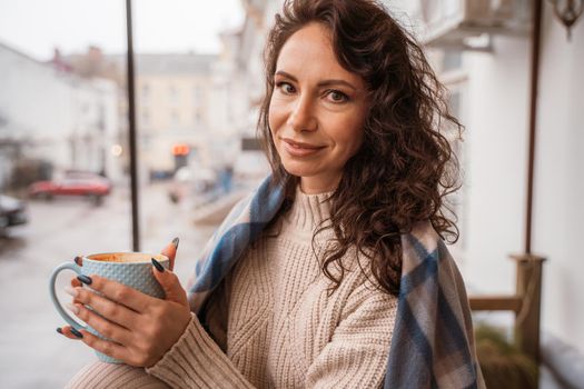 A middle-aged woman in a beige sweater with a blue mug in her hands is in a street cafe on the veranda.