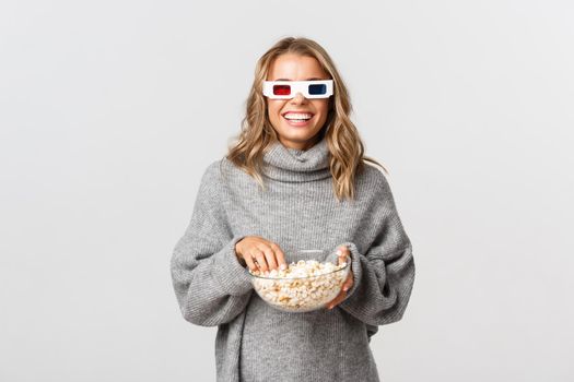 Attractive happy girl watching movie in 3d glasses, eating popcorn, standing over white background.