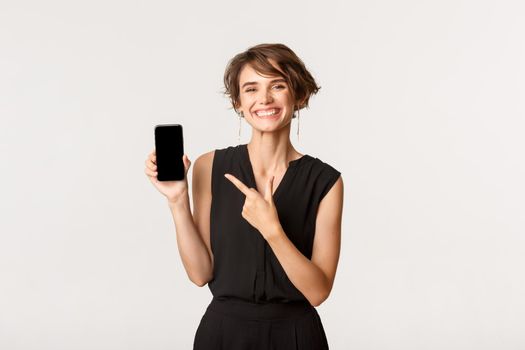 Happy stylish young woman smiling pleased, pointing finger at mobile phone screen, showing application, standing white background.