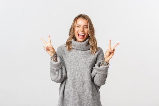 Close-up of attractive blond girl in grey sweater, showing peace signs and smiling, standing over white background.