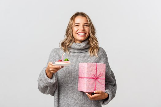 Image of happy blond woman celebrating her birthday, holding b-day cake and gift, standing over white background.