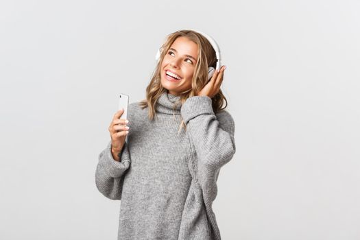 Young attractive blond girl listening music in wireless headphones, smiling and looking at upper left corner, holding mobile phone.