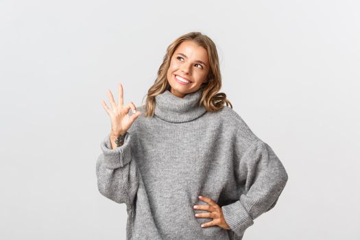 Image of attractive blond woman in casual grey sweater, looking satisfied at upper left corner, showing okay sign in approval, white background.