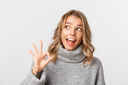 Close-up of excited blond girl in grey sweater, looking amazed left and showing okay sign, standing over white background.