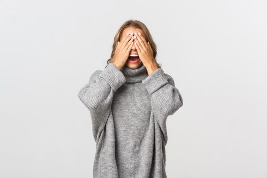 Portrait of beautiful young woman in grey sweater, standing blindsided with hands on face, waiting for surprise, standing over white background.