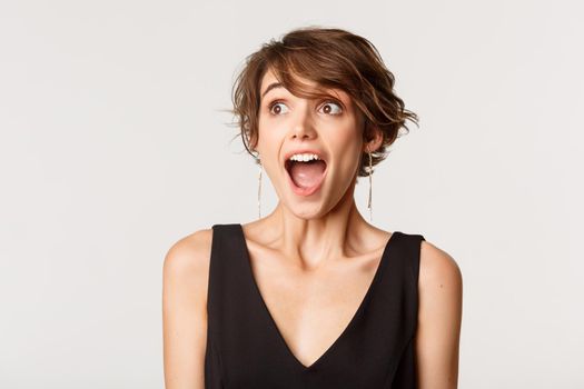 Close-up of excited, amazed young woman drop jaw and looking left, standing over white background.