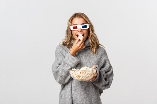 Image of attractive blond girl in 3d glasses, eating popcorn and watching movie, standing over white background.