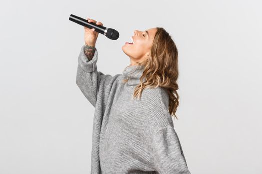Profile shot of happy blond girl singing in microphone. Girl performing a song in karaoke, standing over white background.