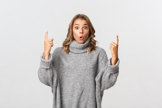 Portrait of surprised blond female in grey sweater, pointing fingers up and looking amazed, standing against white background.