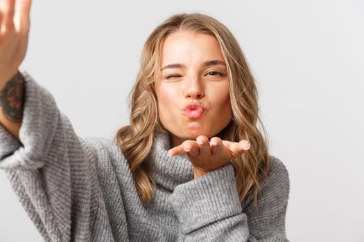 Close-up of attractive blond girl in grey sweater, taking selfie or having video call, sending air kiss at mobile camera, standing over white background.