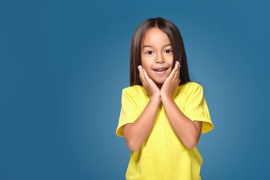 Close up portrait of cute joyful pretty littlegirl with excellent skin and beaming smile, she is admiring her beauty in a mirror, on blue background