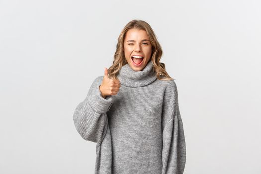 Portrait of attractive happy woman in grey sweater, encourage you, showing thumb-up in approval and winking, recommend good product, like and agree, white background.