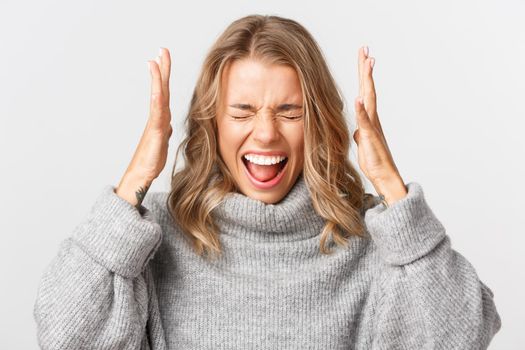 Close-up of angry blond girl in grey sweater, shaking hands distressed, screaming of anger, standing over white background.