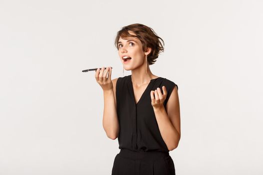 Beautiful girl recording voice message on smartphone, looking up thoughtful, standing white background.