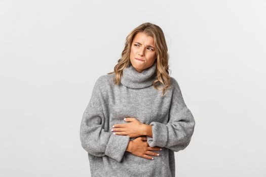 Portrait of blond girl feeling sick, touching stomach and grimacing from pain, have painful menstrual cramps or diarrhea, standing over white background.