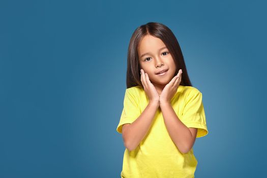 Close up portrait of cute joyful pretty littlegirl with excellent skin and beaming smile, she is admiring her beauty in a mirror, on blue background