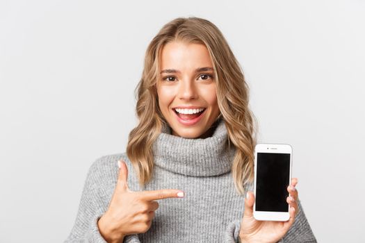 Close-up of attractive blond girl in grey sweater, pointing finger at mobile phone screen, standing over white background.