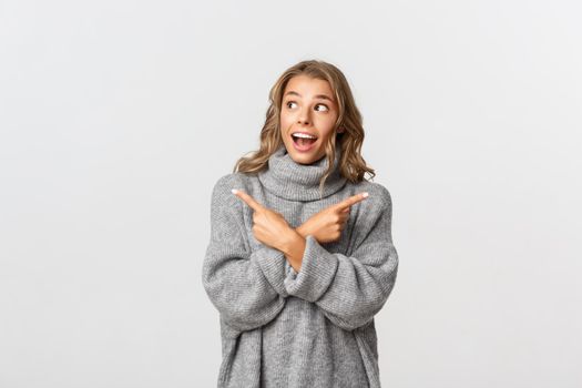 Amazed blond girl pointing fingers sideways, have two choices but looking happy at upper left corner, standing over white background.